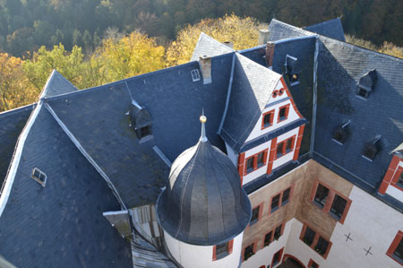 Das Bild zeigt den Ausblick von der Spitze des Bergfriedes aus. Es präsentiert einen Blick über die Dächer von Schloss Rochsburg im Frühling. Im Hintergrund erkennt man den Wald in leichten Grüntönen.