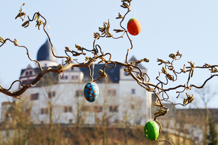 Das Bild zeigt einen Weidezweig mit Ostereiern. Im Hintergrund ist verschwommen Schloss Rochsburg zu sehen.