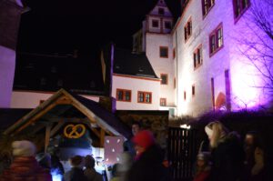 Gästeführer Michael Kreskowsky zeigt einer Gruppe den Backofen im Südzwinger der Rochsburg. Ein Lebkuchen hängt am Ofen. Es ist dunkel. Im Hintergrund ist die Fassade des Schlosses farbig angestrahlt.