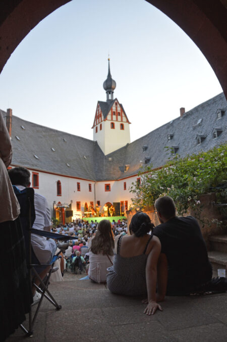 Blick durch den Torbogen in den Wirtschaftshof von Schloss Rochsburg. Im Vordergrund sitzt ein Pärchen auf der Treppe mit dem Rücken zu uns. Der Burghof ist voller Menschen. Am Ende des Hofes ist eine Bühne, auf der gerade eine Band spielt.