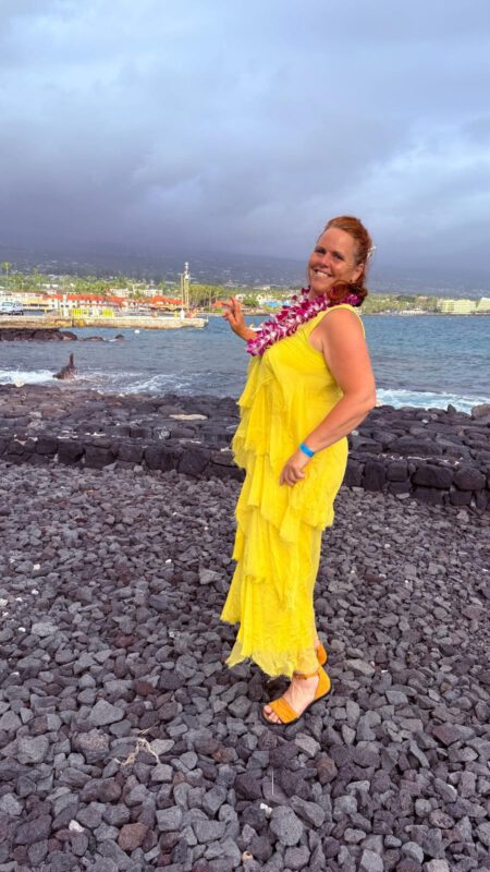 Gästeführerin Carmen Petrus im gelben Kleid und mit Blumenkette, im Hintergrund ein Strand in Hawaii