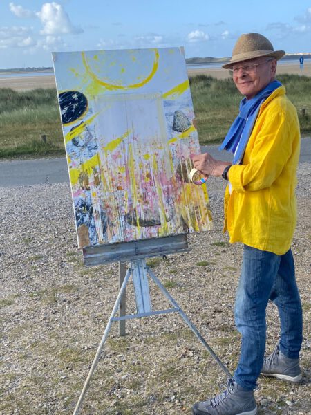 Maler August Ohm mit einer Staffelei, auf der ein neues Kunstwerk entsteht, bunte Farben, im Hintergrund eine Küste