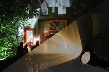 Abendaufnahme des vorderen Burgtores. Es ist dunkel. Ein paar Menschen stehen vor dem Burgtor. Eine Taschenlampe scheint in das Bild.