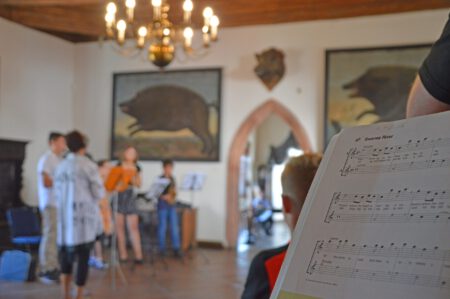 Historischer Saal von Schloss Rochsburg mit Wildschweinporträts, eine Gruppe musizierender Personen, Notenblatt im Vordergrund